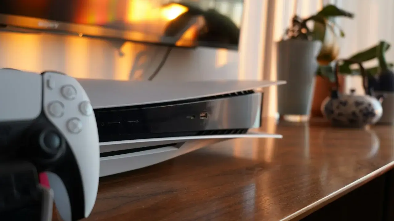 Konsola PS5 i kontroler DualSense na drewnianym blacie, z telewizorem i roślinami w tle.