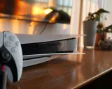 Konsola PS5 i kontroler DualSense na drewnianym blacie, z telewizorem i roślinami w tle.