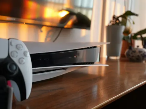 Konsola PS5 i kontroler DualSense na drewnianym blacie, z telewizorem i roślinami w tle.